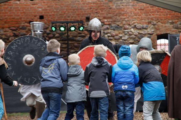Mehrere Kinder stehen vor einem bewaffneten Darsteller mit Schild und Helm bei einem Mittelalterfest vor der Ziegelmauer.