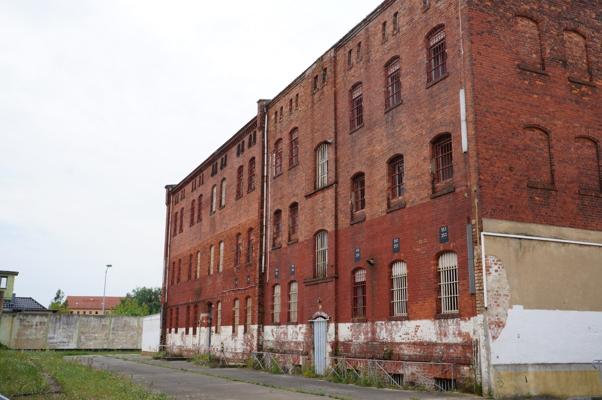 Mehrstöckiges rotes Backsteingebäude mit vielen schmalen Fenstern und verwittertem Sockel neben einer freien Fläche.