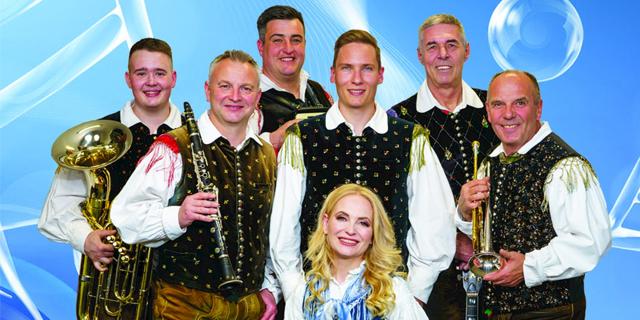 Sechs Musiker und eine blonde Sängerin stehen vor blauem Hintergrund, einige halten Tuba, Klarinette und Trompete.