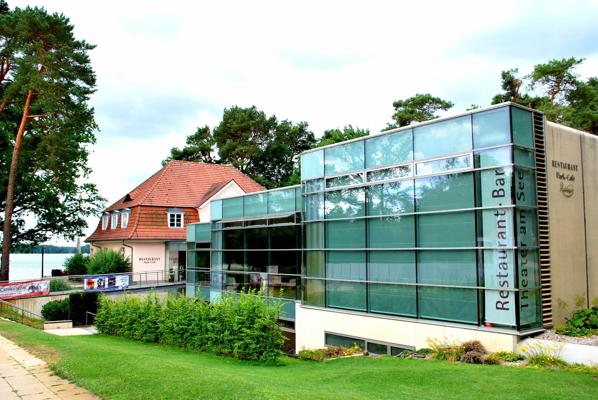 Das Theater am See zeigt eine moderne Glasfassade neben einem roten Gebäude, davor liegt ein gepflegter Rasen am Ufer.