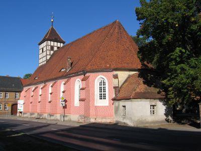 Eine pink verputzte Kirche mit rotem Ziegeldach und Fachwerkturm steht an einer Straße unter blauem Himmel.
