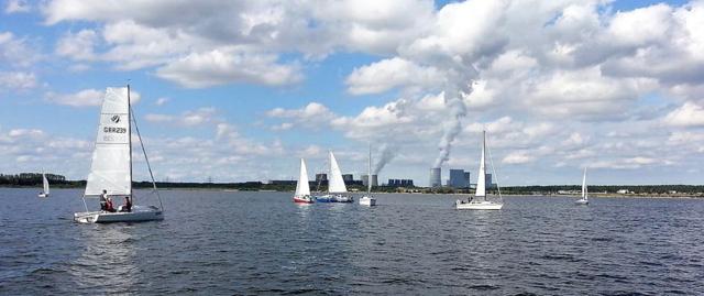 Mehrere Segelboote fahren über einen weiten See, am Horizont stehen Kühltürme und Gebäude unter wolkigem Himmel.
