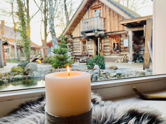 Eine brennende Kerze steht auf der Fensterbank, dahinter sieht man ein rustikales Holzhaus mit Garten und Bäumen im Freien.