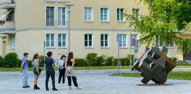 Fünf Personen stehen auf einem Platz und betrachten eine abstrakte Metallskulptur vor einem hellen Wohngebäude mit Bäumen.