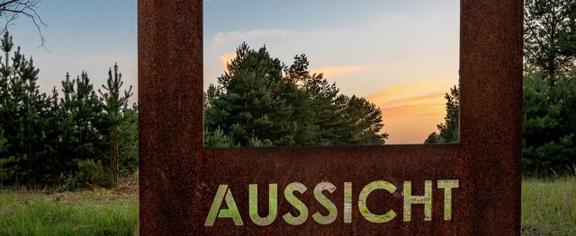 Ein rostiger Stahlrahmen mit dem Schriftzug AUSSICHT steht vor Kiefern und Wiesen im warmen Licht des Sonnenuntergangs.