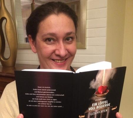 Eine lächelnde Frau hält ein Buch mit schwarzem Cover und dem Titel „Ein Löffel voll begehrte Geschichten“.