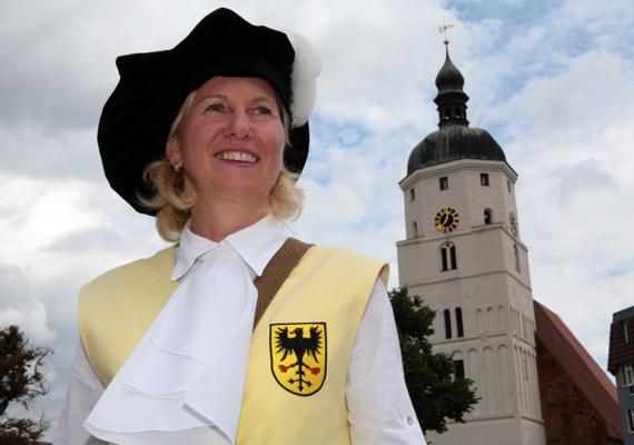 Eine lächelnde Türmerin im historischen Gewand steht vor dem Turm der Paul-Gerhardt-Kirche unter wolkigem Himmel.