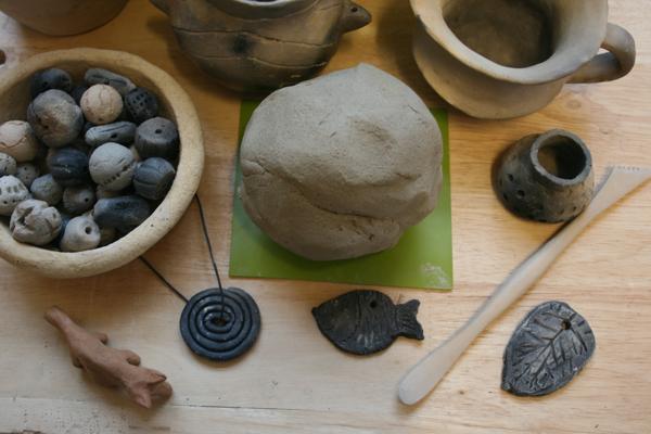 Auf einem Holztisch liegen Schalen, eine Tonkugel, kleine Gefäße, Anhänger und Werkzeuge aus Ton und Keramik.