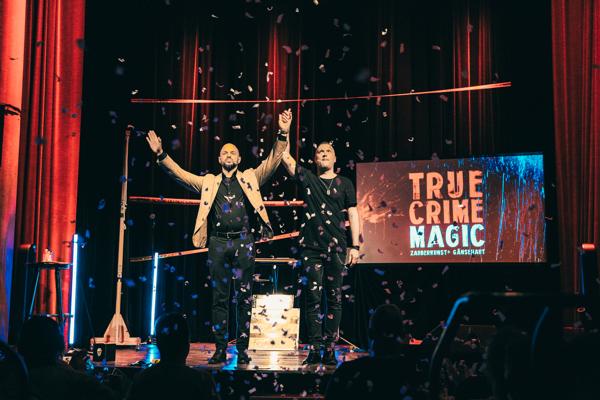 Zwei Männer stehen unter Konfetti auf einer Bühne; neben ihnen leuchtet das Schild „TRUE CRIME MAGIC“.