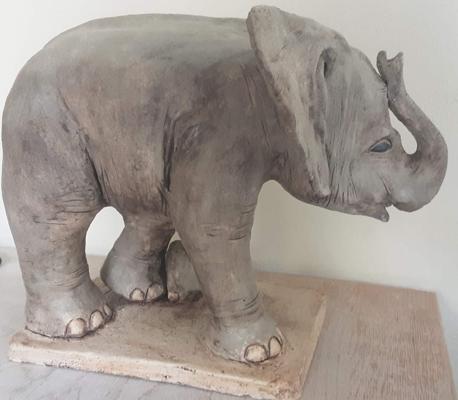 Eine graue Keramikfigur zeigt einen Elefanten mit erhobenem Rüssel auf einer Tonplatte vor hellem Hintergrund.