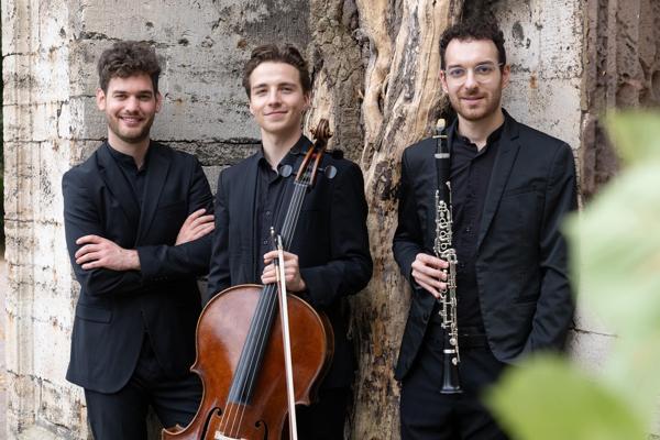 Drei Musiker stehen vor einer alten Steinmauer, einer mit Cello, einer mit Klarinette und einer mit verschränkten Armen.