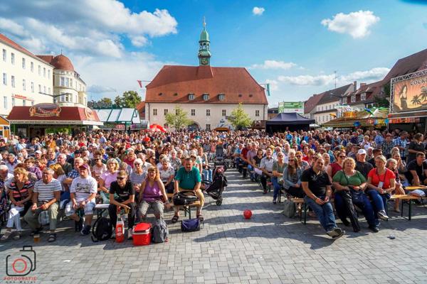 Viele Besucher sitzen dicht gedrängt auf Bänken vor einem historischen Gebäude mit Turmspitze auf dem Marktplatz.