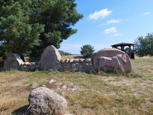Eine steinerne Findlingsmauer mit Tierrelief steht unter einer großen Kiefer auf einer grasigen Fläche.