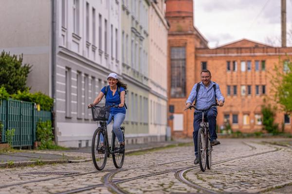 Zwei Radfahrende fahren über ein Kopfsteinpflaster mit Schienen, vorbei an historischen Backsteinbauten.