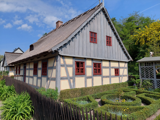 Ein historisches Fachwerkhaus mit Strohdach und roten Fenstern steht neben einer sorgfältig geschnittenen Gartenanlage.