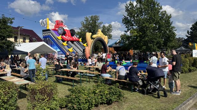 Auf dem Festplatz stehen Bierzeltgarnituren neben einer bunten Riesenrutsche. Viele Besucher sitzen im Sonnenschein.