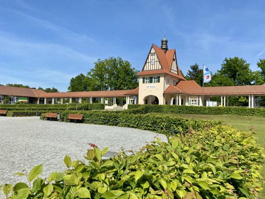 Vor einem Kurhaus mit rotem Dach und Fachwerkturm liegen Hecken, Bänke und eine helle Kiesfläche unter blauem Himmel.