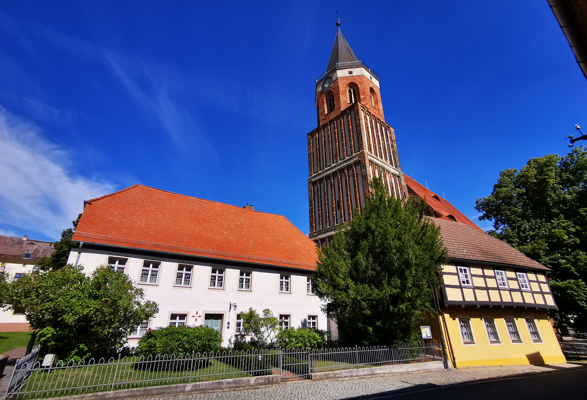 Eine Backsteinkirche mit hohem Turm steht neben weißen und gelben Häusern unter einem klaren blauen Himmel.