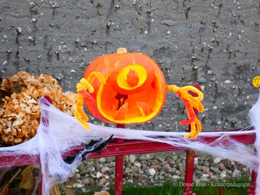 Ein leuchtend bemalter Kürbis mit Spinnenbeinen hängt an einem roten Geländer, umgeben von Spinnennetzen und Herbstlaub.
