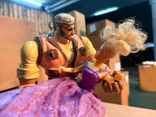 Eine Spielzeugfigur mit Bart und Mütze hält eine Prinzessinnenpuppe im lila Kleid vor Kisten und Holzverkleidung.