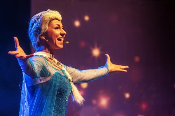 Eine Sängerin im glitzernden blauen Kostüm singt mit ausgebreiteten Armen vor einem warm leuchtenden Bühnenhintergrund.