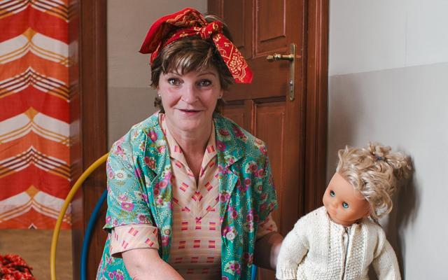 Eine Frau mit rotem Kopftuch und bunter Bluse sitzt in einem Zimmer neben einer blonden Puppe im weißen Pullover.