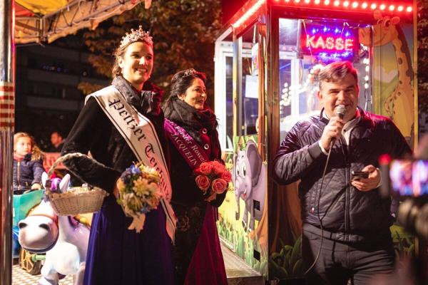 Eine gekrönte Frau mit Schärpe steht neben einer weiteren Frau mit Blumen, während ein Mann am hellen Kassenstand spricht.
