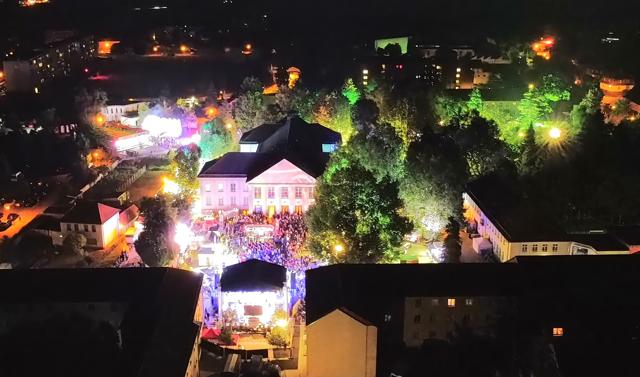 Bei Nacht leuchten ein hell erleuchtetes Kulturhaus, Bühnenlichter und eine große Menschenmenge zwischen den Bäumen.