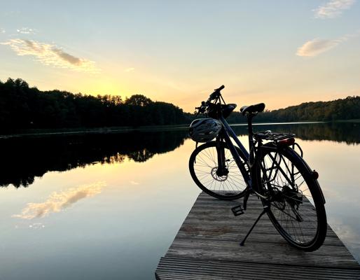 Ein Fahrrad steht auf einem Holzsteg am stillen See, während Himmel und Wolken im warmen Abendlicht im Wasser spiegeln.