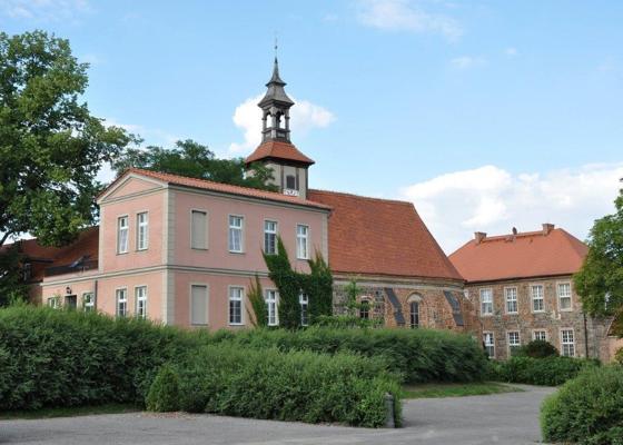 Ein historisches Gebäude mit hellem Putz, rotem Dach und kleinem Turm steht hinter dichten Hecken unter blauem Himmel.