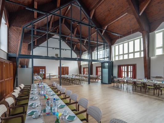In einem großen Saal mit hoher Holzdecke stehen lange Tische mit Tellern, Gläsern und Wasserflaschen entlang der Wände.