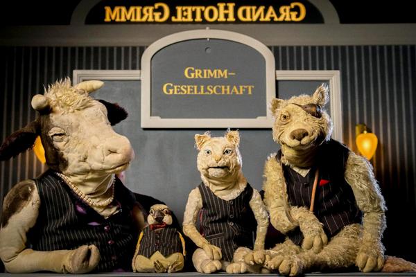 Drei Tierpuppen mit Westen sitzen vor einer Theaterkulisse, darüber steht der Schriftzug „Grimm-Gesellschaft“.