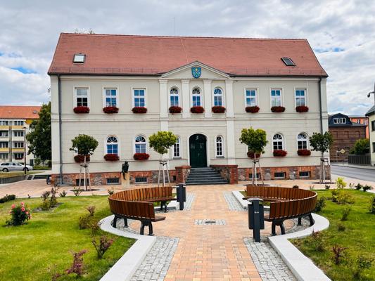 Vor einem hellen Verwaltungsgebäude mit rotem Dach liegen ein gepflegter Garten, runde Sitzbänke und ein gepflasterter Weg.