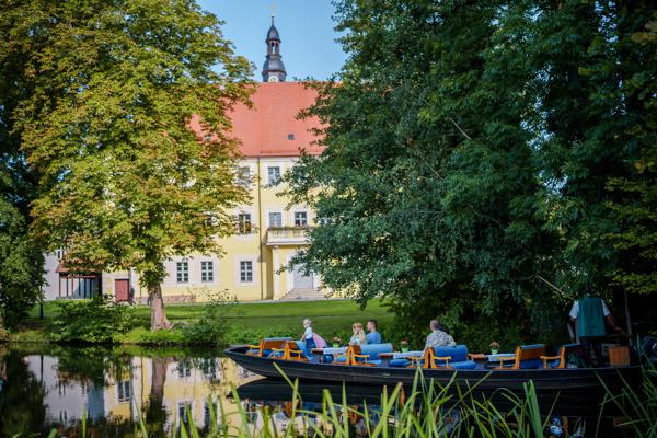 Ein Kahn mit mehreren Fahrgästen gleitet vor einem historischen Schloss am Ufer durch einen stillen Kanal.