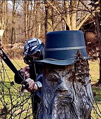 Eine Person mit Schutzhelm zielt beim Arrow Tag mit einem Bogen aus der Deckung hinter einem Baumstamm im Wald.