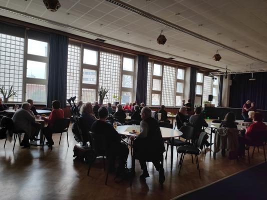 In einem hellen Saal sitzen viele Gäste an runden Tischen, während rechts eine Bühne mit dunklem Vorhang zu sehen ist.