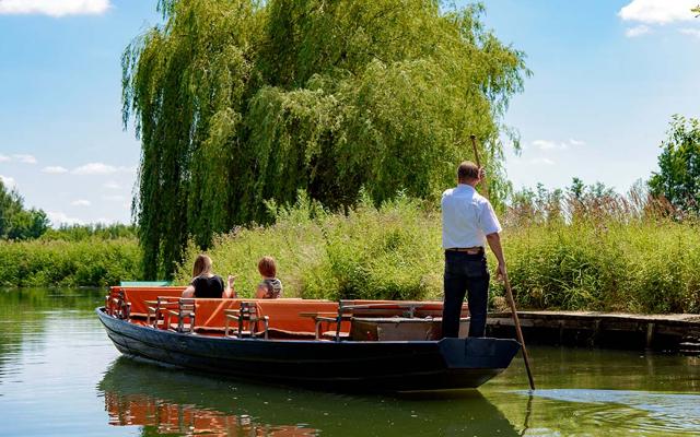 Ein Fährmann stakt einen orangefarbenen Kahn mit zwei sitzenden Gästen auf einem ruhigen Fluss unter einer großen Weide.