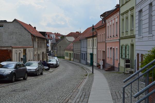 Bunte und schlichte Altstadthäuser säumen eine gepflasterte Straße mit parkenden Autos und Gehweg.