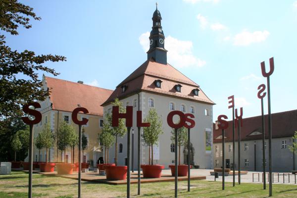 Vor dem Renaissance-Schloss stehen große Buchstaben auf Stangen, dahinter erhebt sich ein heller Bau mit Turm und rotem Dach.