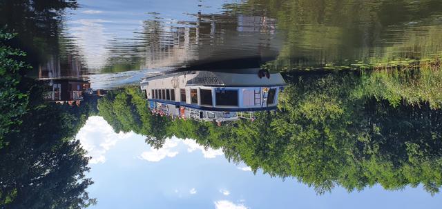Ein Ausflugsschiff liegt auf ruhigem Wasser, umgeben von dichtem Uferwald und deutlichen Spiegelungen im Kanal.