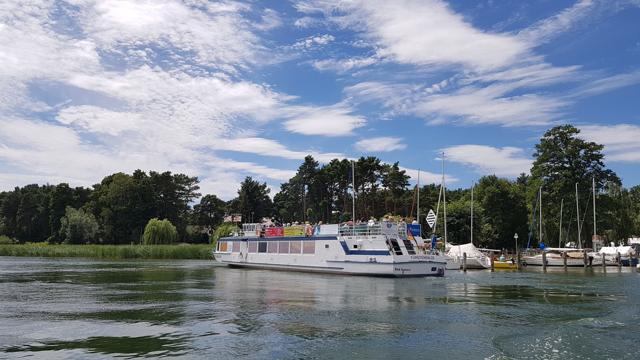 Ein Ausflugsschiff liegt am Ufer vor einem kleinen Hafen mit Segelbooten; dahinter stehen Bäume unter wolkigem Himmel.