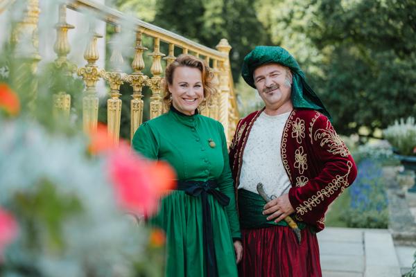 Zwei Personen in historischen Gewändern stehen im Grünen vor einem goldenen Geländer und lächeln in die Kamera.