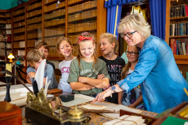 Mehrere Kinder schauen in einem historischen Raum mit Bücherregalen zu einer Frau, die auf ein altes Kräuterbuch zeigt.