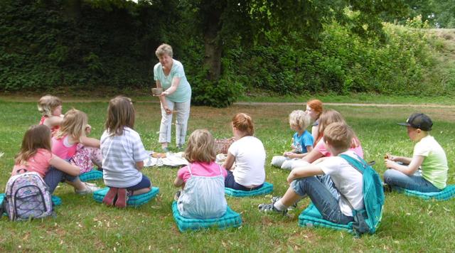 Eine ältere Frau erzählt im Grünen Kindern eine Geschichte, während sie auf Decken und Kissen im Kreis sitzen.