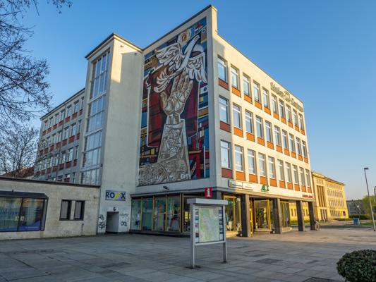 Vor einem modernen Verwaltungsgebäude mit farbigem Wandmosaik liegt ein weiter Platz unter klarem Himmel.