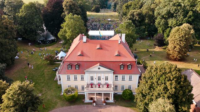 Luftaufnahme eines weiß verputzten Herrenhauses mit rotem Dach, umgeben von alten Bäumen und einer großen Wiese.