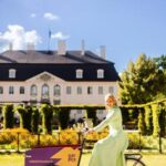 Eine Person in hellem Kleid fährt ein Lastenrad mit violetter Weinkiste vor dem Schloss und einer gepflegten Parkanlage.