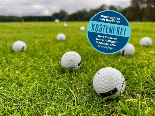 Mehrere Golfbälle liegen auf grünem Rasen, daneben steht ein blaues Schild mit dem Text „Wochenplan mit Kurkarte KOSTENFREI“.