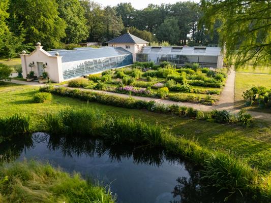 Eine helle Orangerie liegt zwischen gepflegten Beeten, Wegen und einem kleinen Teich am Rand einer Parkanlage.