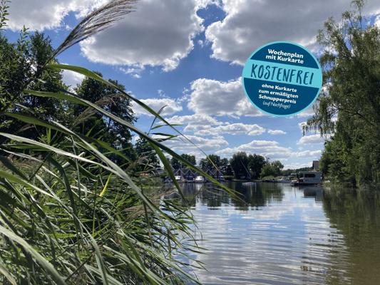 Am ruhigen Ufer mit Schilf und Bäumen steht ein runder Hinweis „Wochenplan mit Kurkarte KOSTENFREI“ vor wolkenreichem Himmel.